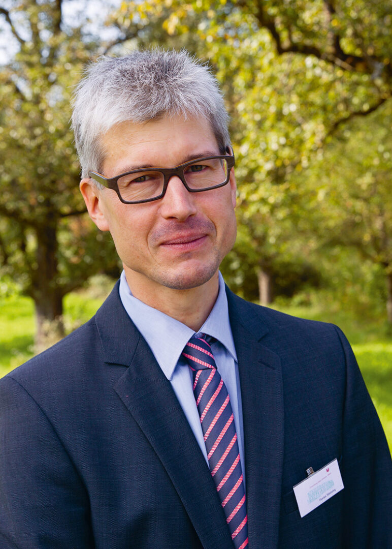 Porträt eines Mannes im Business-Anzug mit gestreifter Krawatte und Brille, aufgenommen im Freien vor grünem Hintergrund mit Bäumen.